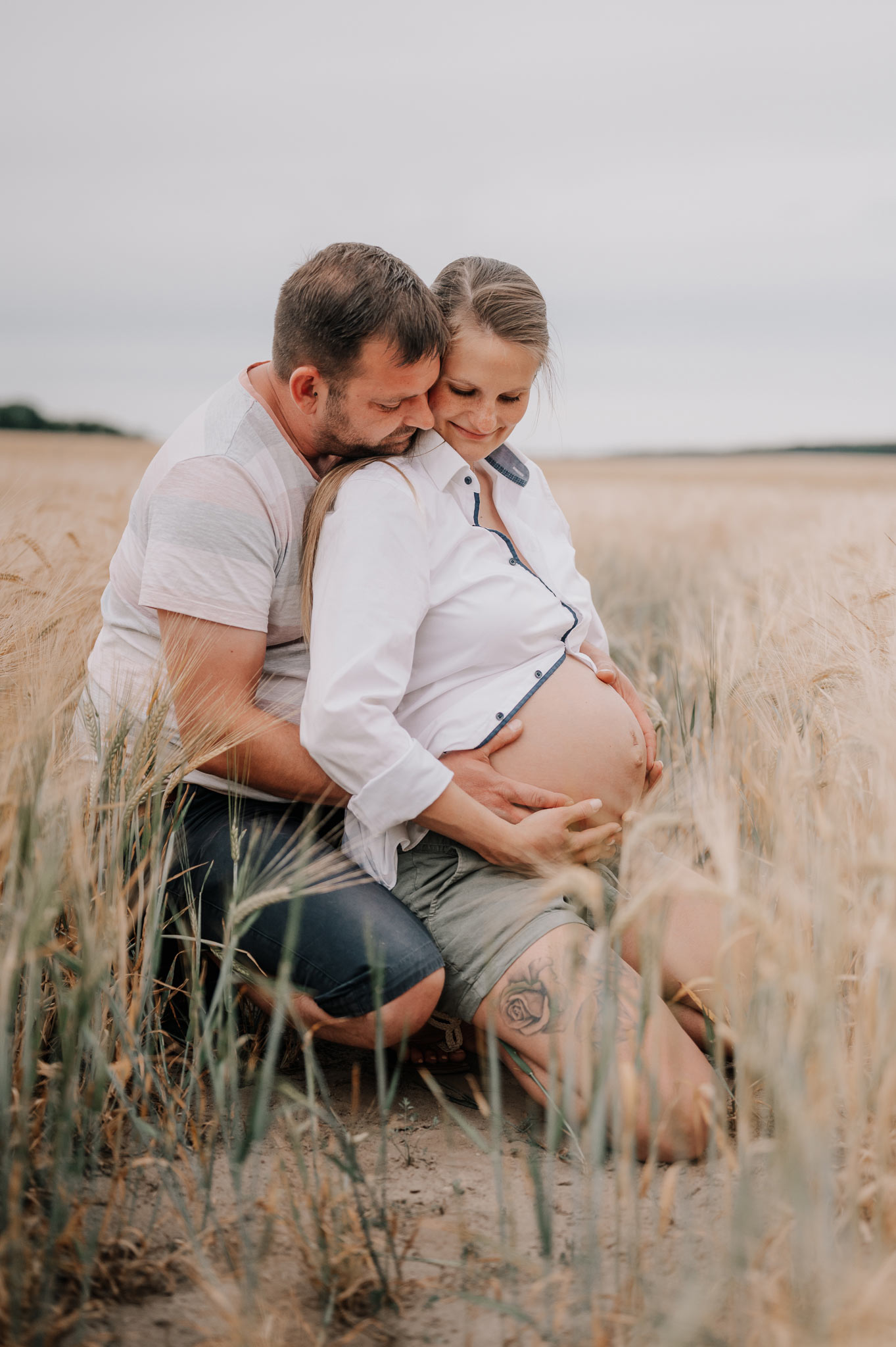 Ein verliebtes Paar kniet in einem Feld. Der Mann streichelt liebevoll den Bauch seiner schwangeren Partnerin.